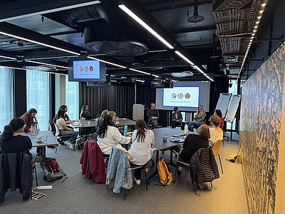 Several people sit at tables in a modern meeting room watching a presentation on screens