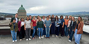 A group of people is standing on a rooftop terrace overlooking the city of Zurich, with a large domed building in the background