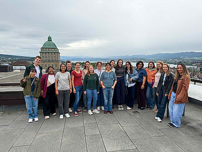 A group of people is standing on a rooftop terrace overlooking the city of Zurich, with a large domed building in the background
