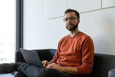 Mann mit rotem Pullover sitzt auf Sofa und arbeitet an Laptop. Es handelt sich um den Forscher Jonas Ricker.