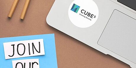 Laptop mit Aufkleber 'CUBE 5 Creating Security', daneben Zettel mit handgeschriebenem Text 'JOIN OUR TEAM', zwei Bleistifte und drei Büroklammern auf braunem Untergrund
