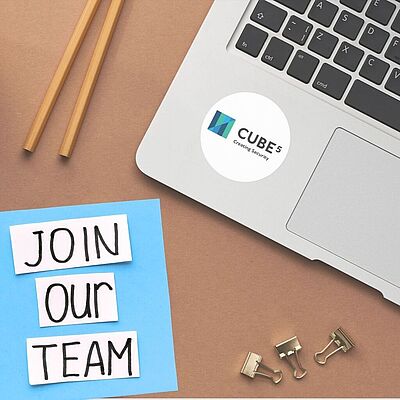 Laptop mit Aufkleber 'CUBE 5 Creating Security', daneben Zettel mit handgeschriebenem Text 'JOIN OUR TEAM', zwei Bleistifte und drei Büroklammern auf braunem Untergrund