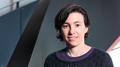 Porträt einer lächelnden Frau mit kurzen dunklen Haaren vor einem dunklen Hintergrund mit diagonalen Linien