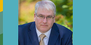 A man in a blue suit in front of a blurred background. It is Michael Bailey.