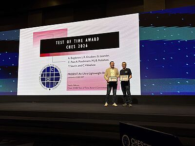 Two men standing on a stage holding certificates in front of a large screen displaying the 'Test of Time Award CHES 2026' with names and a logo