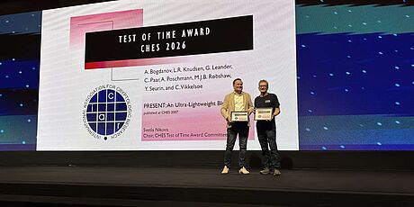 Two men standing on a stage holding certificates in front of a large screen displaying the 'Test of Time Award CHES 2026' with names and a logo