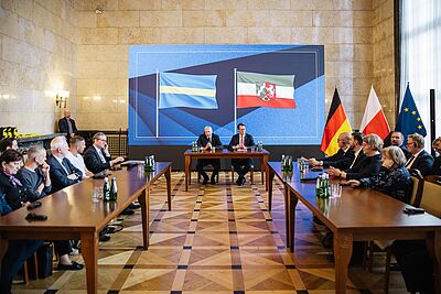 Zwei Männer sitzen an einem Tisch in einem großen Raum mit mehreren Personen an zwei gegenüberstehenden Tischen und Flaggen von Schlesien, Nordrhein-Westfalen, Deutschland, Polen und der EU im Hintergrund.