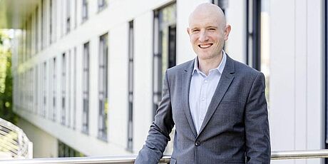 Julian Loss steht im Anzug vor einem Gebäude der Ruhr-Universität Bochum.