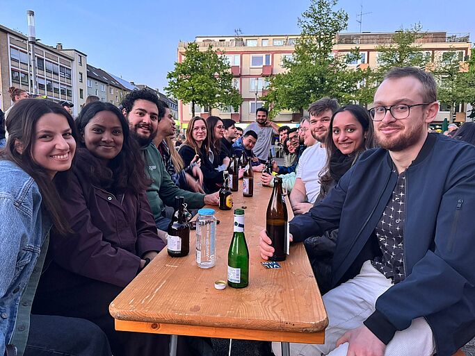 Group of young people sitting at a long wooden table outdoors with drinks smiling at the camera