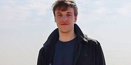 Ein Portrait von CASA PhD Tobias Wienand. Ein Mann mit braunen Haaren steht vor einem hellen Hintergrund.