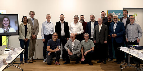 Gruppenfoto von 15 Personen in einem Konferenzraum, einige stehen, andere knien, im Hintergrund ein Bildschirm mit einer weiteren Person und ein Banner mit dem Schriftzug 'CASA'.