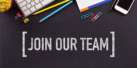 Schwarze Fläche mit der weißen Schrift '[JOIN OUR TEAM]' und verstreuten Büroartikeln wie Tastatur, Stiften, Notizblock, Büroklammern und Smartphone